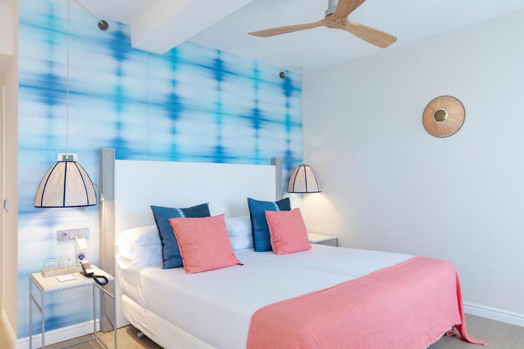 A calm double room at Honucai with a coral and tie-dye-blue headboard, white linen, a slow ceiling fan and a balcony door open to the sea light